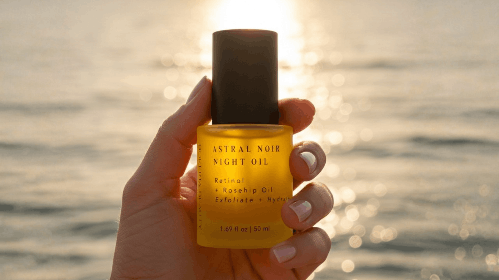 Lady holding bottle of Astral Noir Night Oil in front of the ocean as the sun is setting