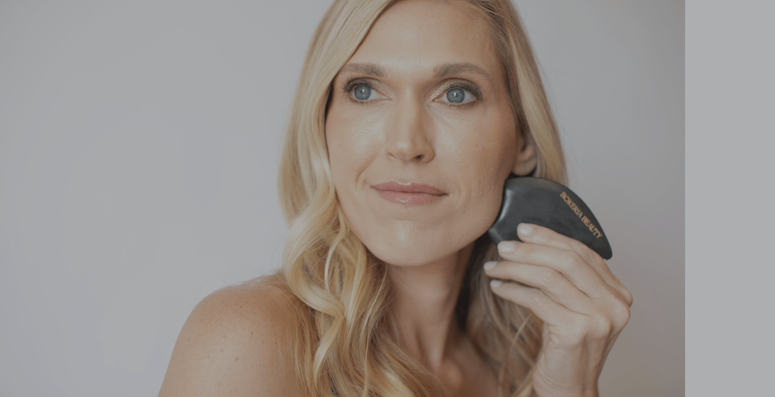 Woman holding a black gua sha tool against her face on a plain background
