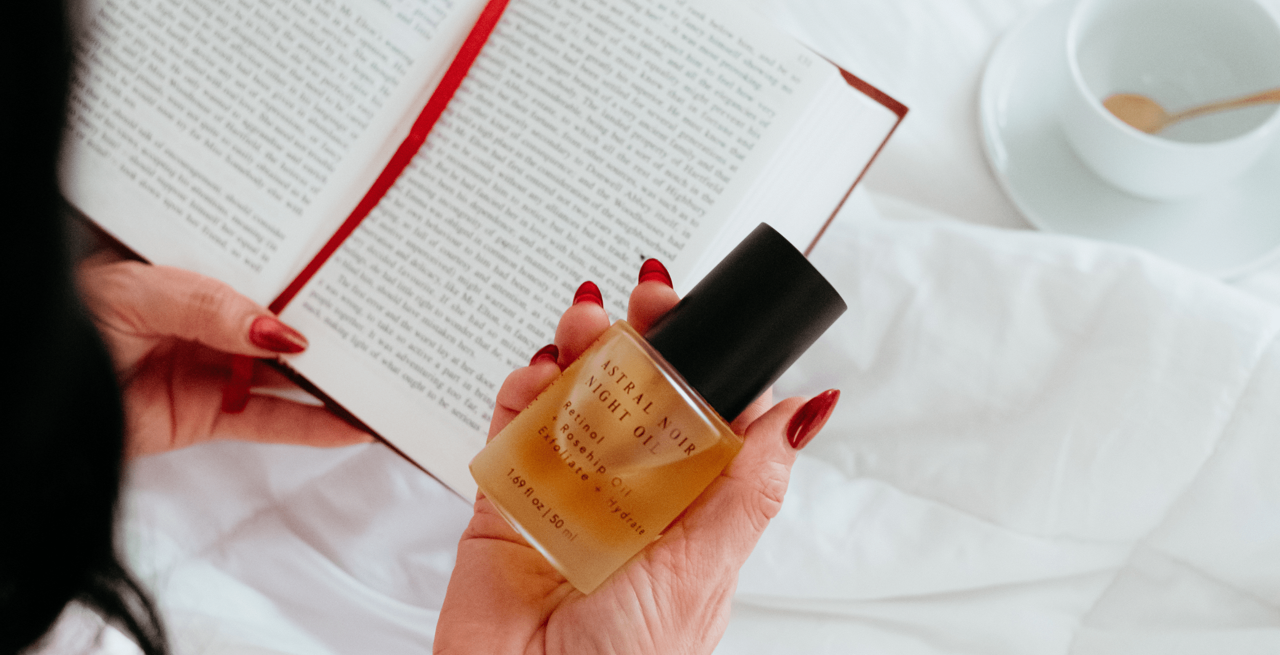 Hand holding a bottle of perfume next to an open book and a cup on a white surface