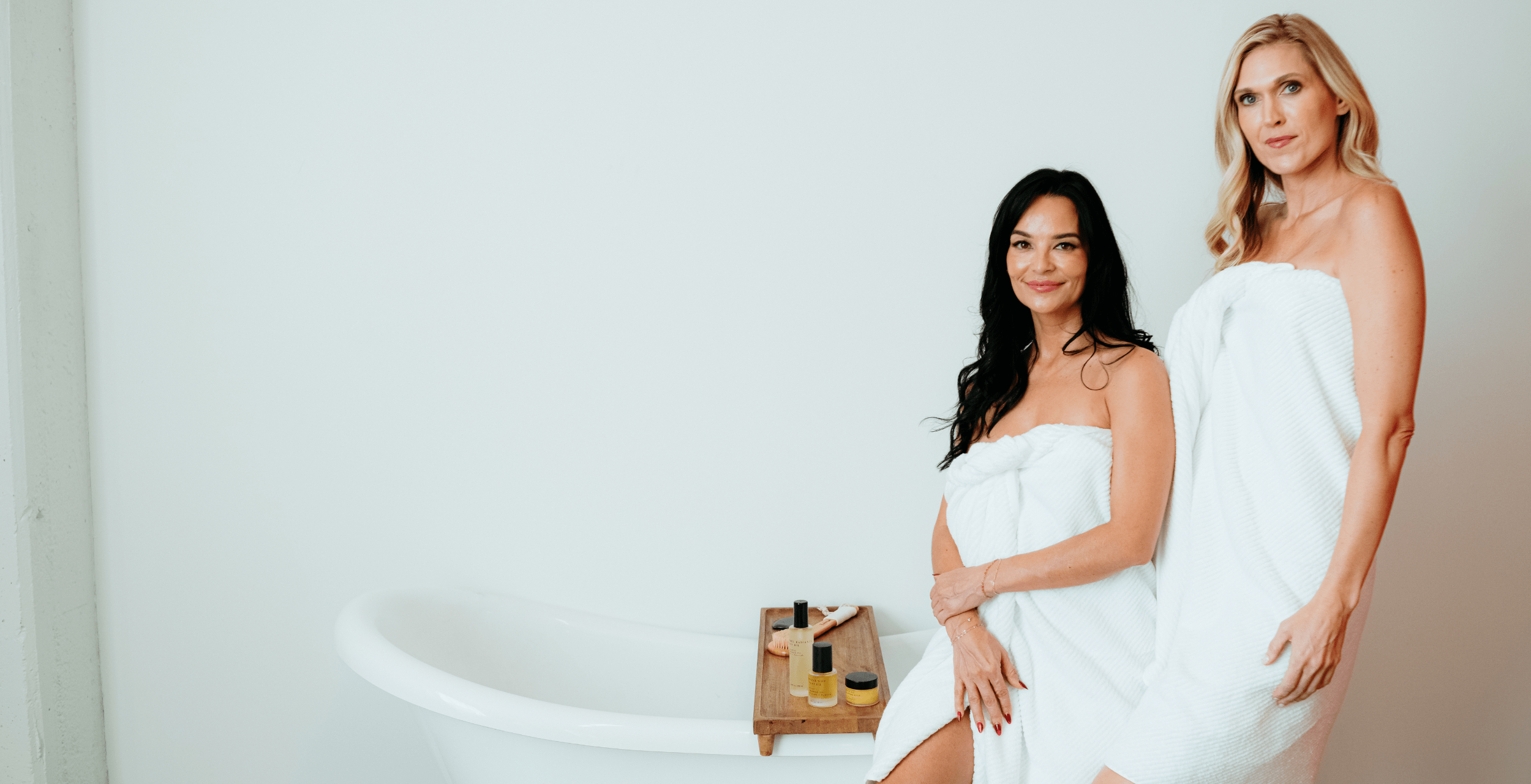 Two women wrapped in white towels standing next to a bathtub with a neutral background