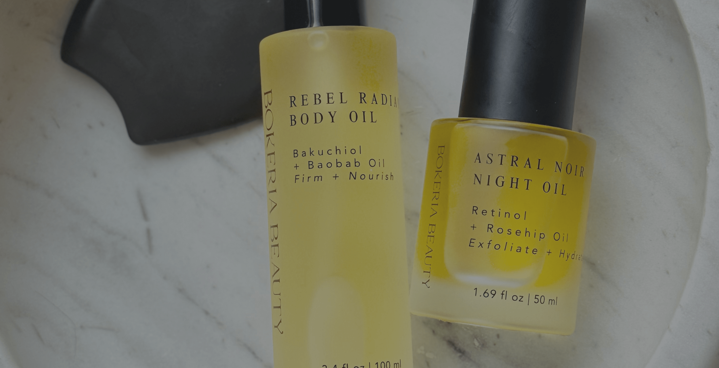 Two yellow bottles of Rebel Radiance body oil and Astral Noir night oil skincare products on a marble surface.