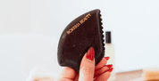 Hand holding a dark brown gua sha tool with 'Bokeria Beauty' branding against a light background